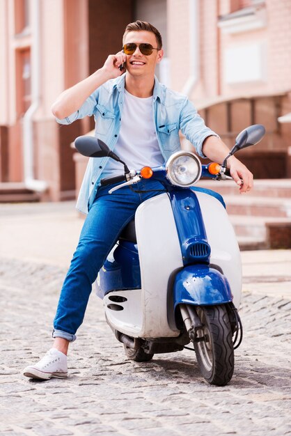 Inviting friends for a ride. Cheerful young man sitting on scooter and talking on the mobile phone