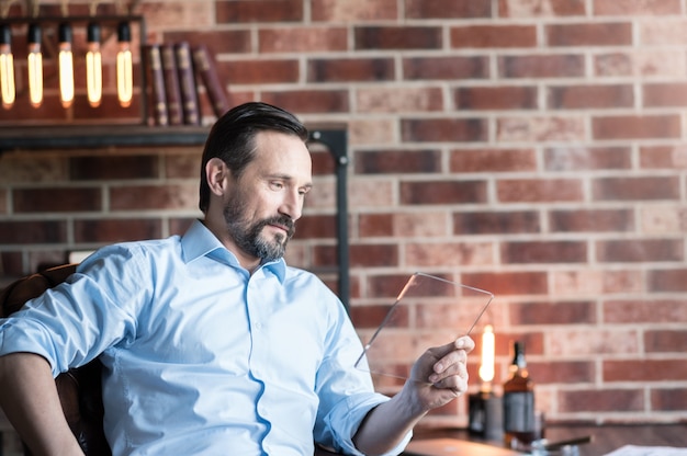 Involved in the activity. Nice handsome bearded man holding a tablet and looking at its screen while sitting in his study