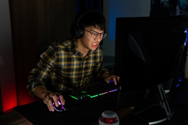 Involved asian man cyber sport gamer concentrated playing video games on computer at night dark room at home, eSport and technology concept.