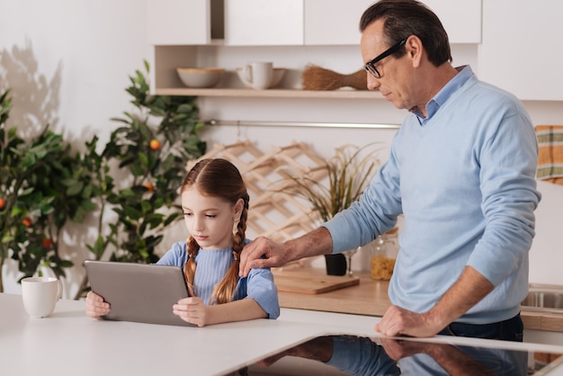 Involved caring aged man standing in the kitchen while expressing dissatisfaction and taking away digital device from little granddaughter