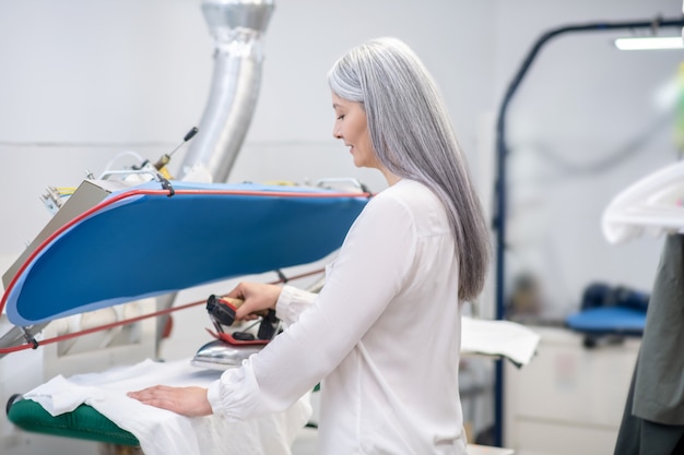 Involved contented woman standing sideways to camera ironing clothes in dry cleaning in good mood