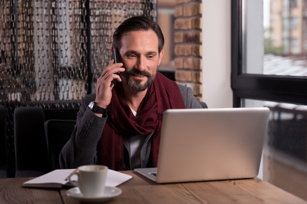 Involved in the conversation. Handsome successful self employed businessman looking at the laptop screen and smiling while speaking on the cell phone