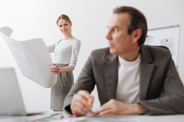 Involved in project. Positive professional female engineer holding a blueprint while her colleague sitting at the table and being involved in thoughts