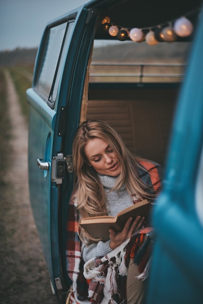 読書に関わっています。青いレトロなスタイルのミニバンの中に座って本を読んで毛布で覆われた魅力的な若い女性