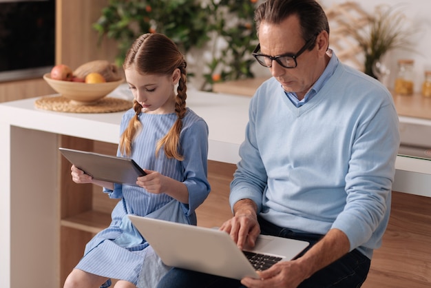 Involved senior retired man sitting at home and using gadgets with granddaughter while surfing the Internet and checking social media profiles