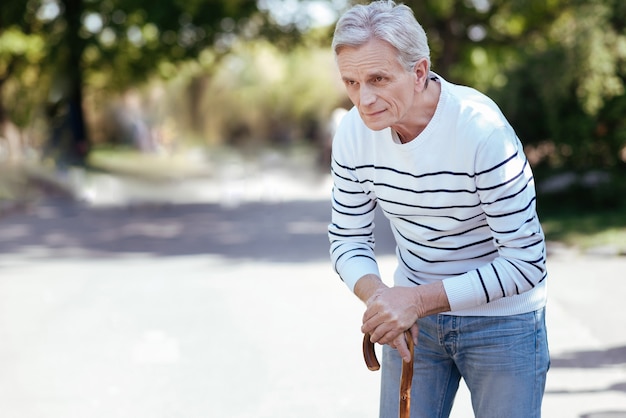 Photo involved thoughtful old man looking away and leaning on the stick while walking in the park