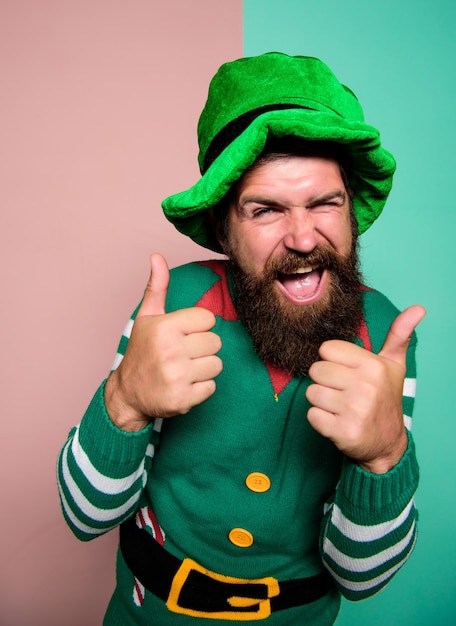 アイルランドのビールの伝統。冬のカーニバル。アイルランドのパブパーティー。聖パトリックの日。緑の帽子で幸せなひげを生やしたヒップスター。クリスマスエルフ。幸せなお祝い。あごひげを生やした陽気な男は、親指を立てて楽しんでいます。