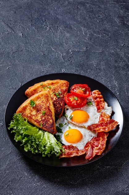 Photo irish breakfast of fried eggs rasher or bacon slices fried potato farls grilled tomatoes and fresh lettuce on black plate on concrete table vertical view free space