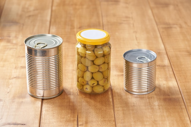 Photo iron tin can with tab opener and olives in a glass jar on the wooden table.