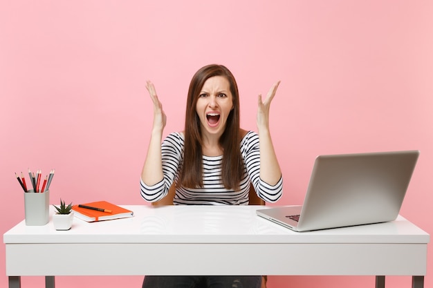 Photo irritated angry woman in casual clothes screaming spreading hand sit work at white desk with contemporary pc laptop isolated on pastel pink background. achievement business career concept. copy space.