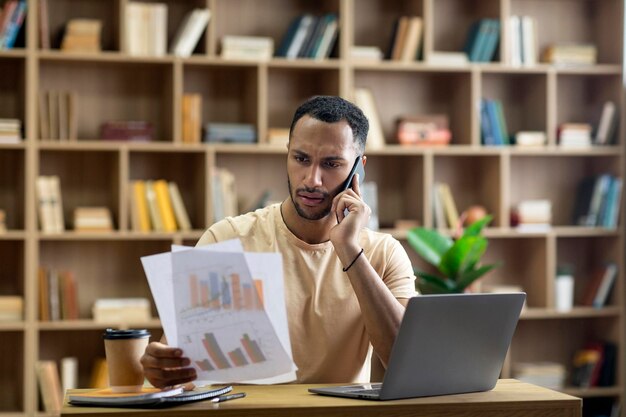 Irritated arab businessman working from home sitting at desk with laptop holding documents and having phone call