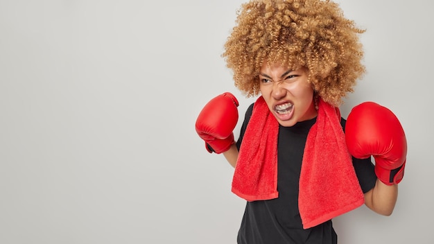 Irritated curly haired active female boxer wears mouthguard and
gloves clenches teeth dressed in black t shirt with red towel
around neck isolated over grey background boxing and sport
concept