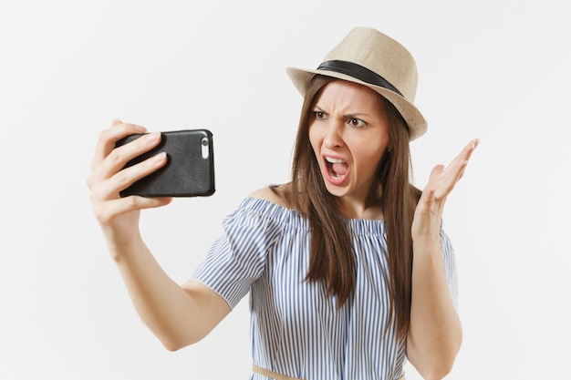 Irritated dissatisfied woman in blue dress, hat doing selfie shot on mobile phone or video call isolated on white background. People, sincere emotions, lifestyle concept. Advertising area. Copy space.