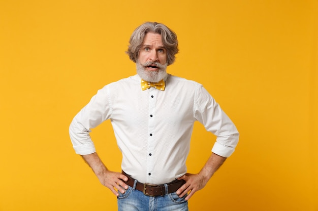 Irritated elderly gray-haired mustache bearded man in white shirt bow tie posing isolated on yellow orange background in studio. people lifestyle concept. mock up copy space. stand with arms akimbo