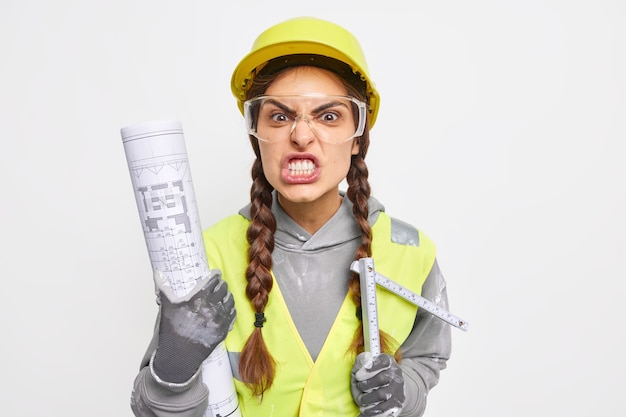 Irritated female builder clenches teeth angrily holds paper blueprint and tape measure going reconstruct dwelling 