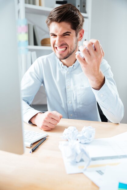 Irritated mad young businessman using computer and crumpling paper on workplace