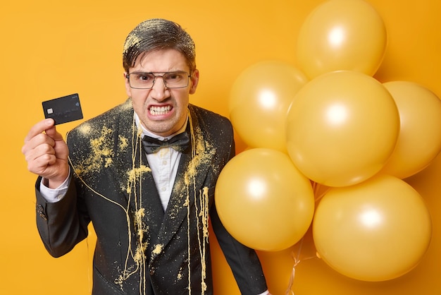 Photo irritated man clenches teeth looks angrily holds banking card and bunch of inflated balloons dressed in festive outfit feels outraged as hasne enough money for celebrtion party time concept