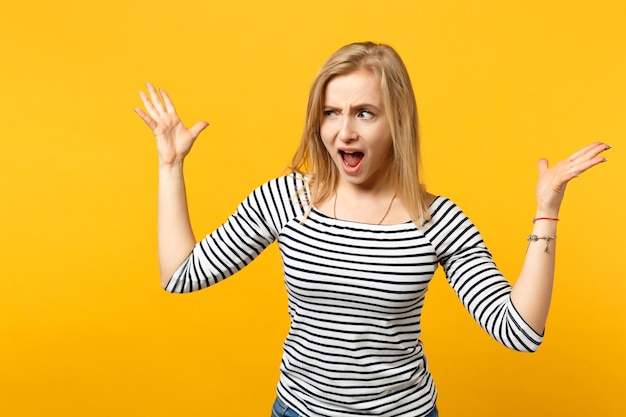 Irritated perplexed young woman in striped clothes looking aside, spreading hands, screaming isolated on yellow orange wall background. People sincere emotions, lifestyle concept. Mock up copy space.