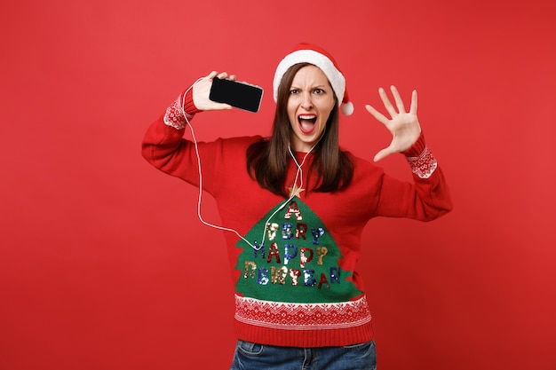 Irritated Santa girl with earphones swearing, spreading hands, hold mobile phone with blank empty screen listen music isolated on red background. Happy New Year 2019 celebration holiday party concept.