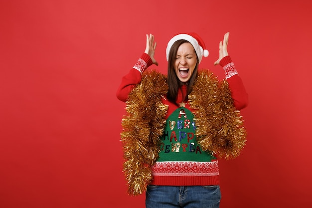 Photo irritated santa girl with golden garland, tinsel spreading hands, keeping eyes closed, screaming isolated on red background. happy new year 2019 celebration holiday party concept. mock up copy space.