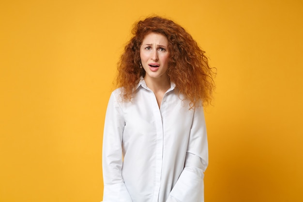 Irritata, scioccata, stanca, giovane donna dai capelli rossi, in camicia bianca casual in posa isolata sul muro giallo arancione orange