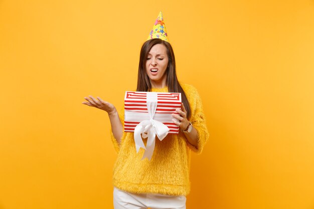 Irritated upset young woman in birthday party hat spreading hands holding red box with gift, present isolated on bright yellow background. People sincere emotions, lifestyle concept. Advertising area.