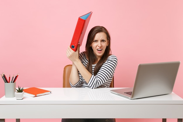 Irritated woman hiding behind red folder with paper document working on project while sit at office with laptop isolated on pastel pink background. Achievement business career concept. Copy space.