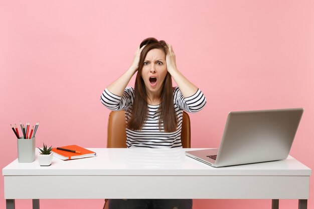 Irritated woman screaming clinging to head sit and work at white desk with contemporary pc laptop isolated on pastel pink background. Achievement business career concept. Copy space for advertisement.