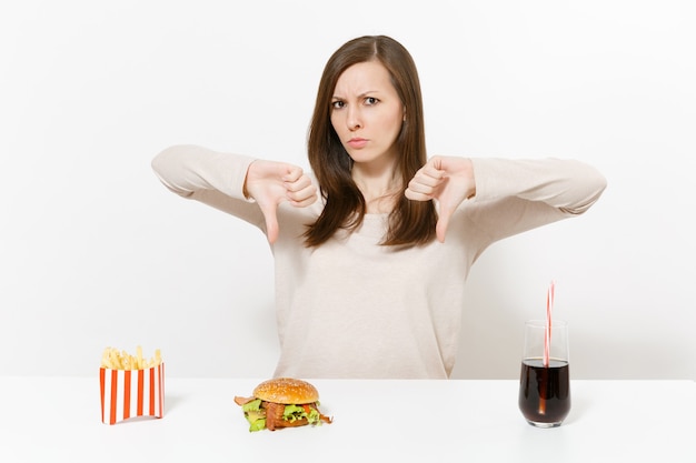 Фото Раздраженная женщина показывает большие пальцы руки вниз за столом с колой картофеля фри гамбургера в стеклянной бутылке изолированной на белой предпосылке. правильное питание или классический американский фастфуд. рекламная площадка с копией пространства.