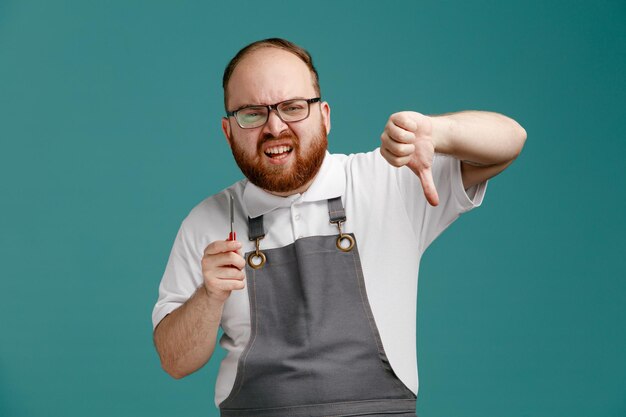 Irritated young barber wearing uniform and glasses holding straight razor looking at camera showing thumb down isolated on blue background