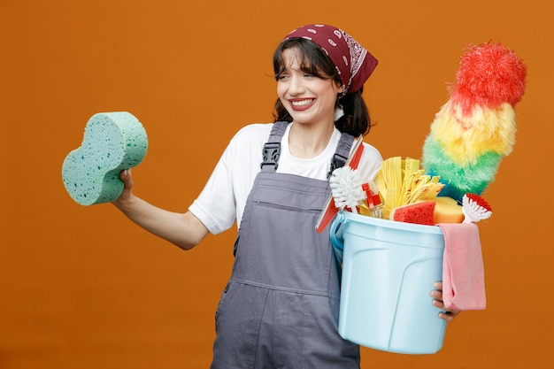 Irritated young female cleaner wearing uniform and bandana holding sponge and bucket of cleaning tools looking at side isolated on orange background