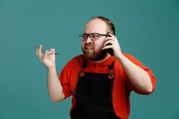 Irritato giovane barbiere maschio con occhiali camicia rossa e grembiule da barbiere che tiene le forbici guardando di lato mentre parla al telefono isolato su sfondo blu