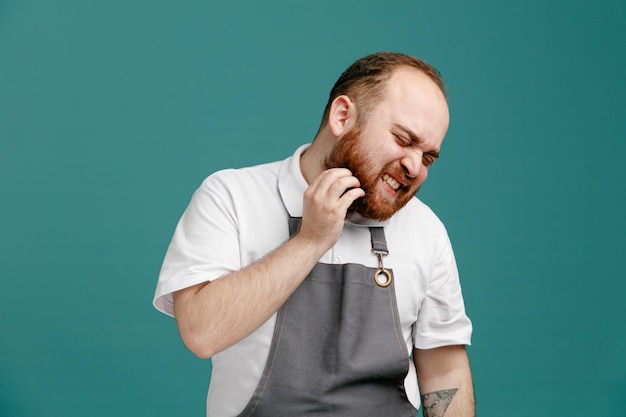 Irritated young male barber wearing white shirt and barber apron turning head to side scratching his beard with eyes closed isolated on blue background