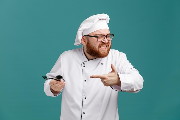 Irritated young male chef wearing glasses uniform and cap holding spoon and fork pointing at them looking at side isolated on blue background