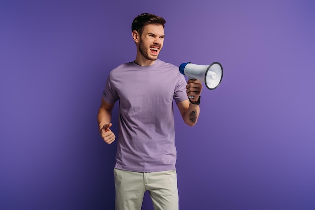 Irritated young man screaming in megaphone while looking away on purple background