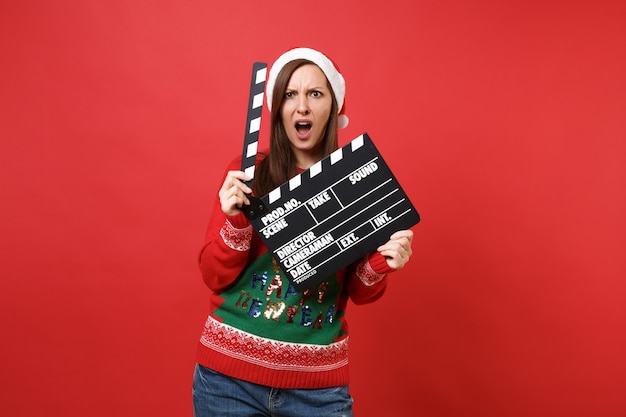 Irritated young Santa girl in Christmas hat swearing, hold classic black film making clapperboard isolated on red background. Happy New Year 2019 celebration holiday party concept. Mock up copy space.