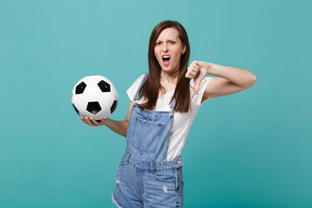 Irritated young woman football fan support favorite team with soccer ball showing thumb down isolated on blue turquoise background. People emotions, sport family leisure concept. Mock up copy space.