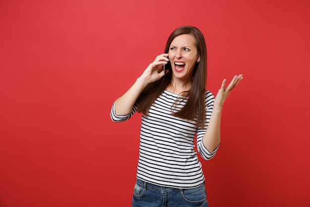 Irritated young woman screaming spreading hands talking on mobile phone, conducting conversation isolated on bright red wall background. People sincere emotions, lifestyle concept. Mock up copy space.