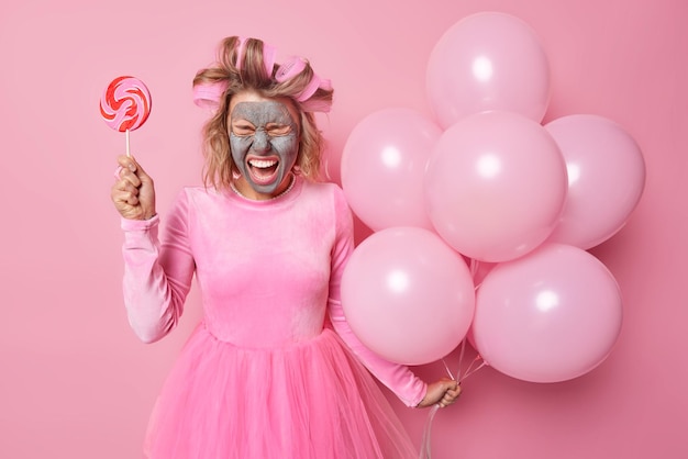 Раздраженная молодая женщина кричит от раздражения, держит кучу воздушных шаров, карамельные конфеты накладывают бигуди для волос, а косметическая маска носит платье, готовится к празднованию на розовом фоне