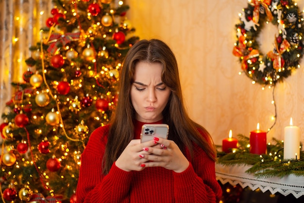 Giovane donna irritata con lo smartphone in mano che invia messaggi di testo con gli amici bellissime luci dell'albero di natale e candele sullo sfondo