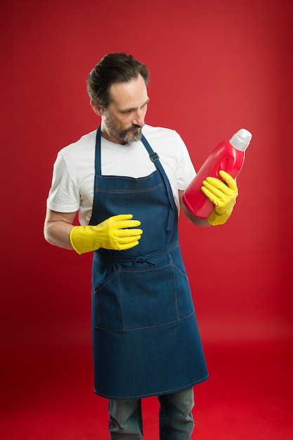 Is this detergent good for my clothes. Eldery household worker with laundry detergent in rubber gloves. Household laundering. Senior man ready for doing laundry. Mature man holding detergent bottle.