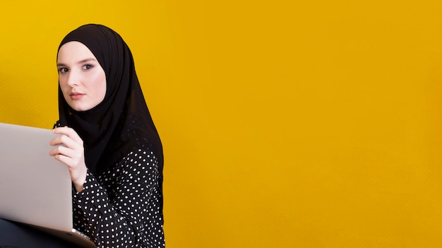 Donna islamica che esamina il computer portatile della tenuta della macchina fotografica sopra il contesto giallo luminoso