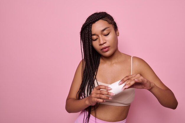 Isolated close-up beauty portrait on pink background of African woman with dreadlocks in beige underwear holding antiperspirant in her hands. Purity, hygiene, body care concept. Copy space