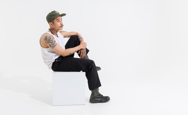 Photo isolated cutout studio shot asian vintage classy mustache with neck arms hands tattoos male fashion model in casual fashionable sleeveless shirt cap boots sitting on square box on white background
