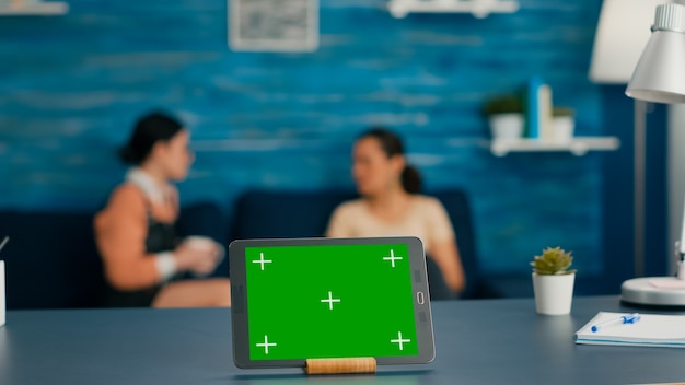 Isolated digital tablet with mock up green screen chroma key display standing on office desk in living room. In background room collegues talking about online communication
