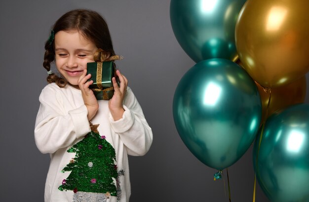 Isolated portrait on gray background of adorable Caucasian little girl with closed eyes gently hugging her Christmas gift, standing near beautiful shiny air balls of green metallic and golden colors