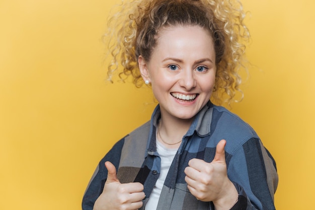 Photo isolated shot of beautiful happy young female with attractive appearance raises two thumbs dressed in casual checkered shirt has curly bushy pony tail isolated over yellow studio background