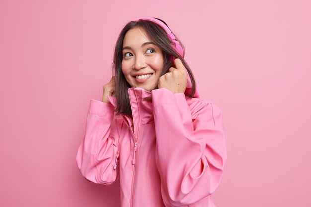 Isolated shot of happy teenage music fan wears wireless headphones on ears smiles broadly dressed in stylish jacket has walk during spare time isolated over pink wall. Hobby and lifestyle