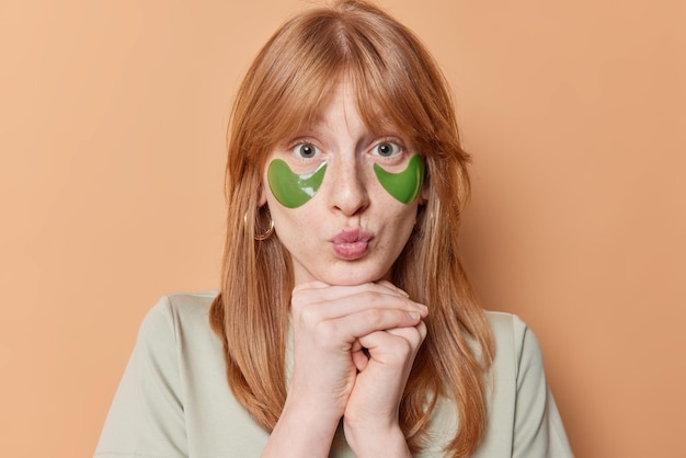 Isolated shot of surprised young ginger female model keeps lips rounded hands under chin puts on green hydrogel patches under eyes dressed casually isolated over brown background Beauty concept