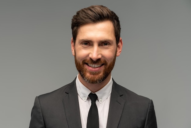 Isolated shot of young handsome male with beard and trendy hairdo wearing formal clothes posing with toothy smile in studio against grey background